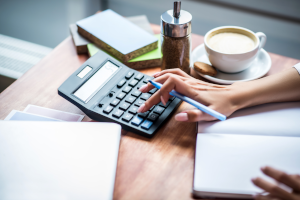 Photo of a person's hand using a calculator at a desk, with a cup of coffee.
